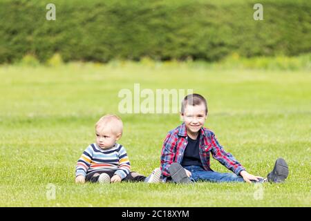 Zwei fröhliche Jungen Brüder verschiedener Altersgruppen spielen Spaß, sitzen auf dem Gras an einem warmen Sommertag. Das Konzept der Freundschaft der Brüder und glückliche Kindheit Stockfoto