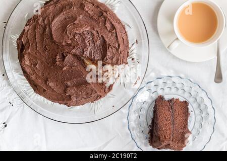 Schokoladenkuchen und Scheibe auf Glas Servierplatte und Tasse Tee auf weißem Tischtuch - Draufsicht Bild Stockfoto