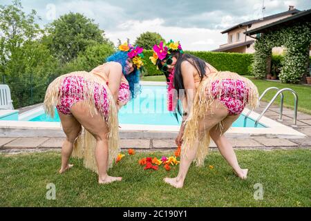 Zwei Freundinnen in hawaiianischen Kostümen pflücken Blumen von einer Wiese vor dem Swimmingpool Stockfoto