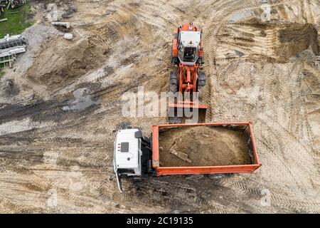 Ein Bagger lädt Sand auf einer Baustelle in einen LKW. Draufsicht von einer Drohne. Stockfoto