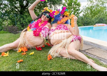 Zwei Freundinnen in hawaiianischen Kostümen haben Spaß auf einer Wiese vor dem Swimmingpool zu sitzen Stockfoto