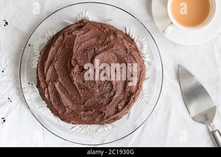 Schokoladenkuchen ganz auf Glas Servierplatte, Metall-Server und Tasse Tee auf weißem Tischtuch - Top V Stockfoto