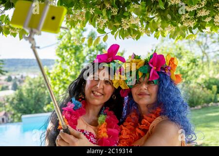 Zwei lächelnde Freundinnen in hawaiianischen Kostümen machen ein Selfie mit einem Smartphone am Pool Stockfoto