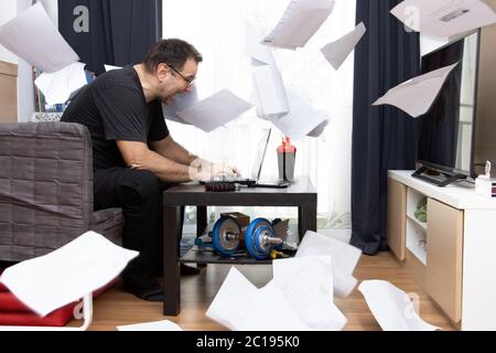 Ein dynamischer Eifer Mann sitzt im Wohnzimmer und arbeitet auf Laptop, Papierdokumente fliegen herum. Ein emotionales Home Office. Stockfoto