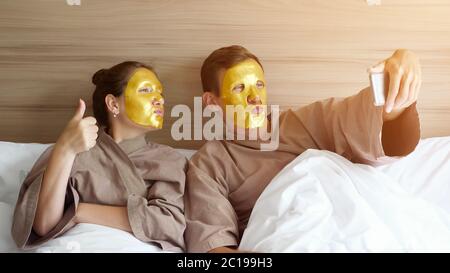 Junge Familie in Bademänteln mit goldenen Gesichtsmasken macht Selfie mit Smartphone im weichen Bett im komfortablen Hotelzimmer liegend Stockfoto