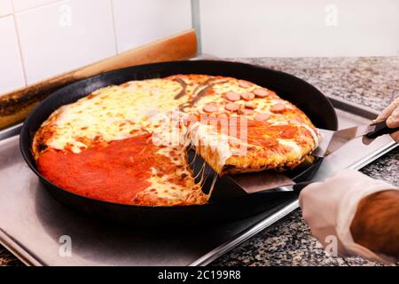 Der Koch nimmt eine Scheibe frisch gebackene Pizza aus einem Backblech und verwendet dabei einen Spatel aus nächster Nähe auf seine Handschlöcke Stockfoto