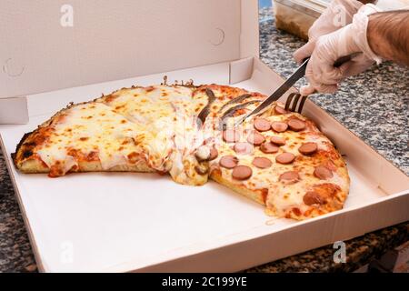 Koch mit Handschuhen schneidet eine Pizza mit verschiedenen Belägen in eine Pappschachtel zum Mitnehmen auf einer Granittheke in Nahaufnahme Stockfoto