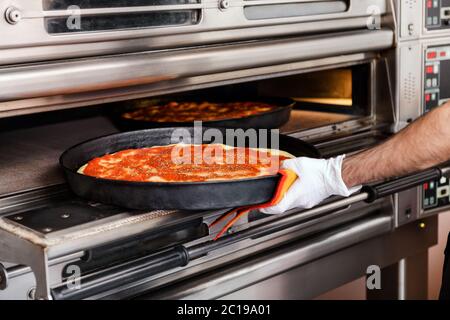 Koch platziert eine Pizza auf einem Backblech in einen Ofen in einer Pizzeria oder Restaurant in einer Nahaufnahme auf seine Hand und das Essen Stockfoto