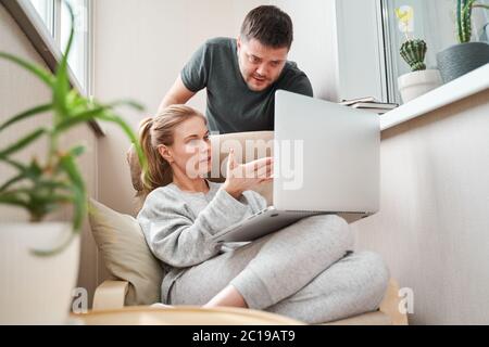 Junge Frau sitzt auf beigefarbenem Sessel mit Laptop und Mann In der Wohnung in der Nähe von ihr stehen Stockfoto