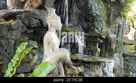 Schöne weiße Stein meditierende Buddha-Statue in der Nähe von künstlichen Wasserfall auf Felsen mit grünem Gras und kleinen Tempel Modell Stockfoto