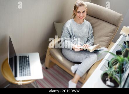 Junge Frau mit Buch in den Händen sitzt auf beige Sessel in der Wohnung Stockfoto