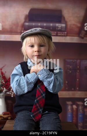 Sweet Kleinkind Junge, gekleidet smart casual, sitzen auf einem Holzstuhl, spielen mit Spielzeug, Regale mit Büchern hinter ihm Stockfoto