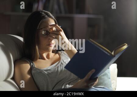 Frau mit Schmerzen leiden Augenbelastung Lesen eines Buches sitzt auf dem Sofa in der Nacht zu Hause Stockfoto