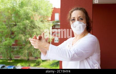 Porträt einer alten Frau in einer medizinischen Maske, die vom Balkon aus applaudiert, in Dankbarkeit gegenüber den Gesundheitshelfern. Einengung durch Coronavirus. Covid-19-Konep Stockfoto