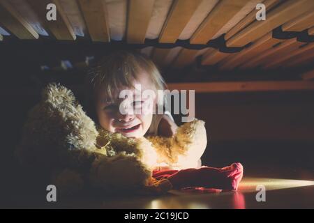 Kleines Kind, versteckt unter dem Bett, umarmt Teddybär und hält Taschenlampe, erschrocken und traurig Stockfoto