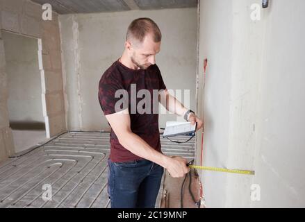 Seitenansicht eines ernsthaften Mannes im Hemd, der Notebook hält und Maßband in der Küche mit Fußbodenheizungsrohren verwendet. Handwerker, der Messung während der Arbeit an der Renovierung der Wohnung. Stockfoto