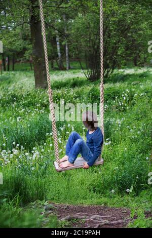 Süßes Kind, vorjugendlicher Junge, schwingend auf einer hölzernen Schaukel auf einem Baum nahe Löwenzahnfeld Stockfoto