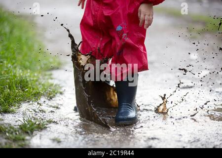 Süßer kleiner Kleinkind Junge, blondes Kind in rotem Regenmantel und blauen Stiefeln, im Regen in schlammigen Pfützen spielen, springen und lachen Stockfoto