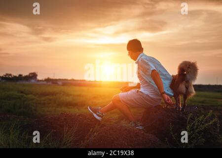 Ein Mann und ein Hund spielen zusammen auf dem Stein bei Sonnenuntergang. Stockfoto