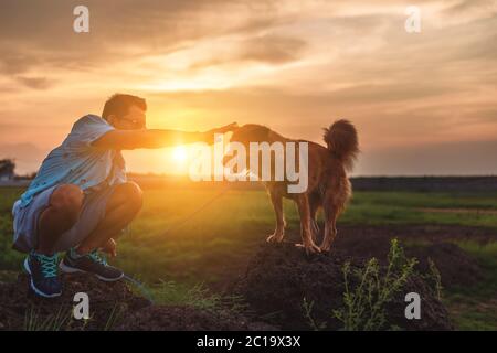 Ein Mann und ein Hund spielen zusammen auf dem Stein bei Sonnenuntergang. Stockfoto