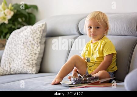 Süßer Kleinkind Junge, sitzt auf einer Couch, essen Kirschen und Blick auf Bilderbuch, genießen gesunde Mahlzeit Stockfoto