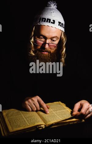 chassidischer jude beim Lesen der Tora. Religiöser orthodoxer jude mit Seitengelenken und rotem Bart in weißem Ballen, der im Dunkeln betet. Low-Key-Foto Stockfoto