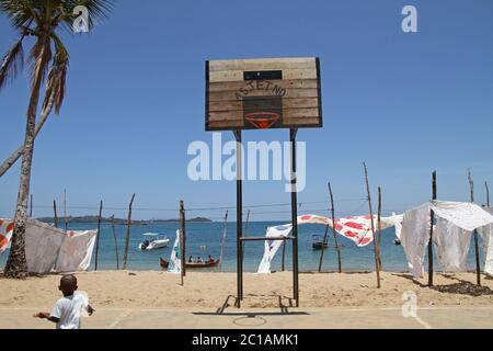 Tischdecken zum Verkauf hängend zum Verkauf vor dem Basketballplatz am Strand mit Booten, Ampangorinana Village, Nosy Komba Island, Madagaskar. Stockfoto