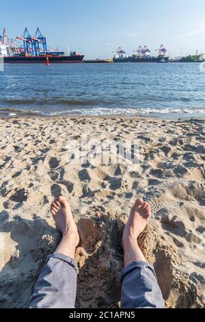 Entspannen mit Füßen im Sand am Elbestrand in Hamburg mit Containerhafen im Hintergrund Stockfoto