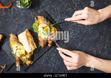 Weibliche Hände hält ein Sandwich mit Eiern Benedikt, Speck und Salat auf schwarzem Stein Hintergrund. Draufsicht Stockfoto