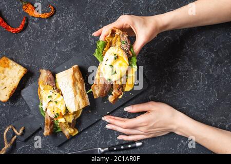 Weibliche Hände hält ein Sandwich mit Eiern Benedikt, Speck und Salat auf schwarzem Stein Hintergrund. Draufsicht Stockfoto