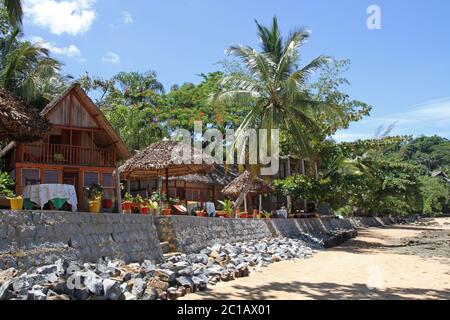 Wand unter Strohdach Lapas und Strandhäuser am Strand, Ampangorinana Village, Nosy Komba Island, Madagaskar. Stockfoto