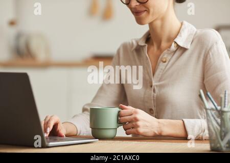 Beschnittenes Porträt einer lächelnden jungen Frau, die Kaffee trinkt, während sie einen Laptop am gemütlichen Arbeitsplatz im Büro benutzt, Kopierraum Stockfoto