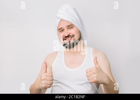 Glücklich lächelnder kaukasischer bärtiger Mann in whilte T-Shirt und mit einem Handtuch auf dem Kopf posiert vor der Kamera auf isoliert, während Hintergrund und zeigt seine Stockfoto