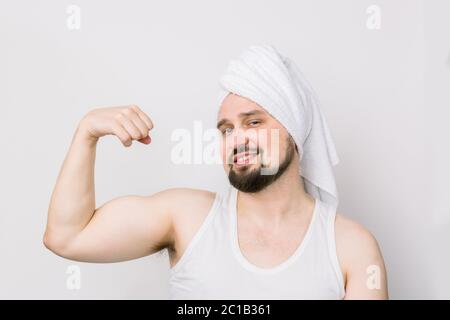 Lustiger junger Kaukasischer mit Schnurrbart und Bart, mit weißem Handtuch auf dem Kopf, lächelnd an der Kamera, zeigt seinen Bizeps. Körper oder Haut Pflege für die Gesundheit des Menschen Stockfoto