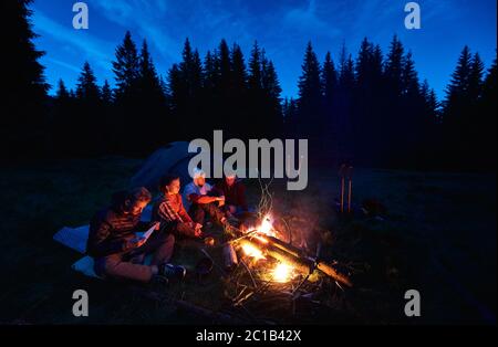 Abend Camping in der Nähe von Feuer, Wald auf dem Hintergrund. Gruppe von vier Freunden Wanderer sitzen zusammen am Lagerfeuer, genießen frische Luft in der Nähe Zelt mit Touristen Ausrüstung in der Nähe, dunkel entspannende Atmosphäre. Stockfoto