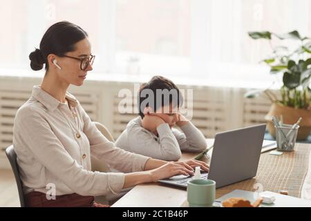 Warm getönte Seitenansicht Porträt der jungen Mutter im Home Office mit Sohn mit Smartphone neben ihr in gemütlichen Küche Innenraum, kopieren Raum Stockfoto