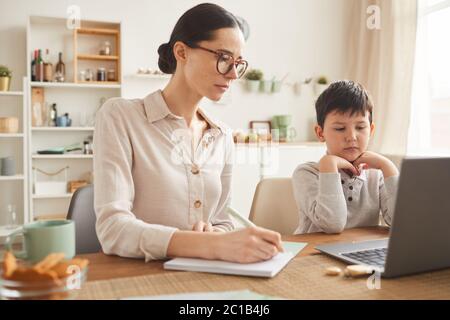 In warmen Farbtönen gehaltene Darstellung der jungen Mutter, die dem Sohn hilft, online zu studieren, während er am Schreibtisch sitzt und einen Laptop in einer gemütlichen Wohneinrichtung verwendet Stockfoto