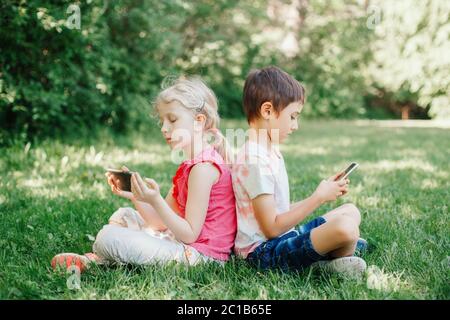 Junge und Mädchen spielen Spiele auf Smartphones im Freien. Kinder digitale Gadget Bildschirm sucht. Kinder Freunde spielen online sitzen auf Gras im Park. Stockfoto