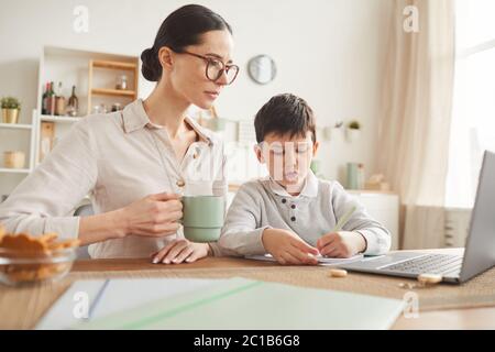 In warmen Farben gehaltene Porträt einer jungen Mutter, die dem Sohn hilft, online zu studieren, während er am Schreibtisch sitzt und einen Laptop in einer gemütlichen Wohneinrichtung verwendet, in der er Platz für Kopien hat Stockfoto