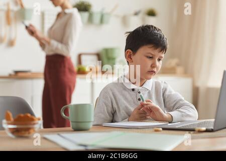 In warmen Farbtönen gehaltene Porträt eines Jungen, der zu Hause studiert, während er einen Laptop am Schreibtisch in einer gemütlichen Wohneinrichtung mit Kopierfläche verwendet Stockfoto