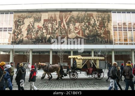 DRESDEN, DEUTSCHLAND - 8. DEZEMBER 2012: Fiaker mit Pferdeantrieb, der in einer Straße voller Touristen vor einem Wandmosaik der DDR-kommunistischen Propaganda auf t Stockfoto