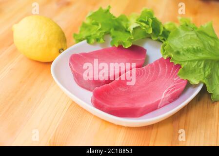 Zwei Stücke rohes frisches Thunfischfilet auf einem Teller mit großen Blättern grünen Salats und gelbe Zitrone auf einem Holztisch. Köstliche SE Stockfoto