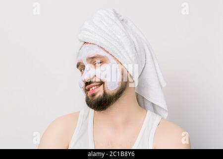 Porträt eines schönen fröhlich entspannten jungen bärtigen Mann mit einer Gesichtsschlamm-Ton-Maske, mit Handtuch auf dem Kopf, posiert mit lustigen Emotionen auf isoliert Stockfoto