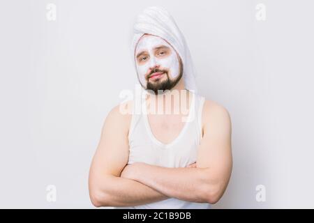 Schöner bärtiger Mann mit weißem Handtuch auf dem Kopf und kosmetischer Maske auf dem Gesicht, steht mit gekreuzten Armen auf weißem Hintergrund. Verfahren zur Kosmetologie Stockfoto