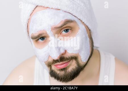 Nahaufnahme des Gesichts eines hübschen, seriösen bärtigen Mannes mit weißem Handtuch auf dem Kopf und einer Schlammreinigungsmaske, der auf weiß isoliert auf die Kamera schaut. Hautpflege Stockfoto