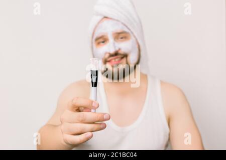 Jouful stattlich bärtigen Mann Anwendung Gesicht Schlammmaske auf Gesicht, zeigt kosmetische Bürste an der Kamera, isoliert auf weißem Hintergrund, Studio-Aufnahme. Konzentrieren Sie sich auf Stockfoto