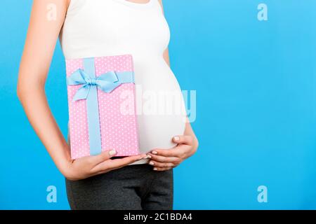 Zugeschnittenes Bild von rosa gepunkteten Geschenkbox in der Hand der schwangeren Frau gegen ihren Bauch auf blauem Hintergrund. Zukünftige Mutter in Heimkleidung. Warten auf einen BA Stockfoto