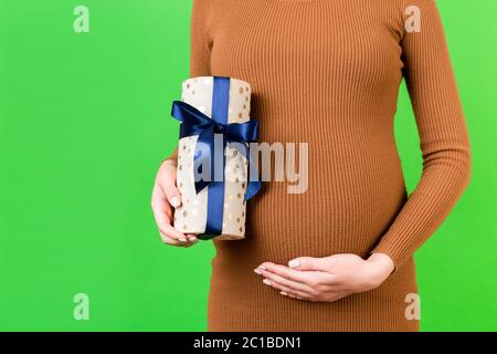 Zugeschnittenes Bild von schwangeren Frau in braunem Kleid mit einer Geschenkbox und berühren ihren Bauch auf grünem Hintergrund. Erwartet einen kleinen Jungen. Speicherplatz kopieren. Stockfoto