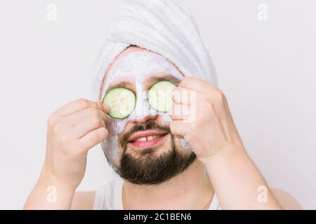 Schöner unrasiert Mann mit weißem Handtuch auf dem Kopf und Ton Maske auf der Haut, bedeckt die Augen mit zwei Scheiben Gurke, posiert auf weißem Hintergrund Stockfoto