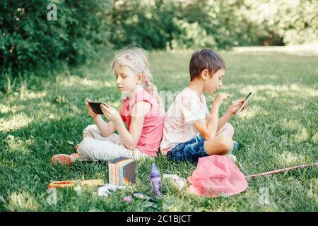 Junge und Mädchen spielen Spiele auf Smartphones im Freien. Kinder digitale Gadget Bildschirm sucht. Kinder Freunde spielen online sitzen auf Gras im Park. Stockfoto